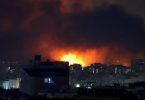 Noćni snimak grada s velikim eksplozijama i vatrom na horizontu, dim i plamen prekrivaju небо