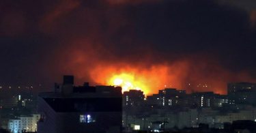 Noćni snimak grada s velikim eksplozijama i vatrom na horizontu, dim i plamen prekrivaju небо