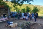 Grupa ljudi okupljena oko ruševne građevine mlinice u prirodnoj sredini s brdima u pozadini
