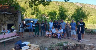 Grupa ljudi okupljena oko ruševne građevine mlinice u prirodnoj sredini s brdima u pozadini