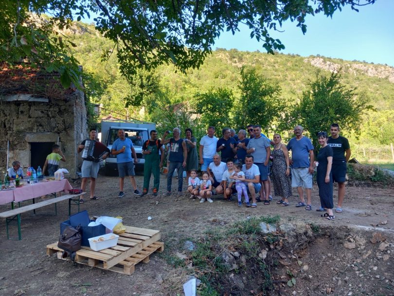 Grupa ljudi okupljena oko ruševne građevine mlinice u prirodnoj sredini s brdima u pozadini