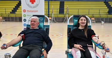 Dva dobrovoljca sjedeće tijekom darivanja krvi u sportskoj dvorani, s logom Crvenog križa u pozadini