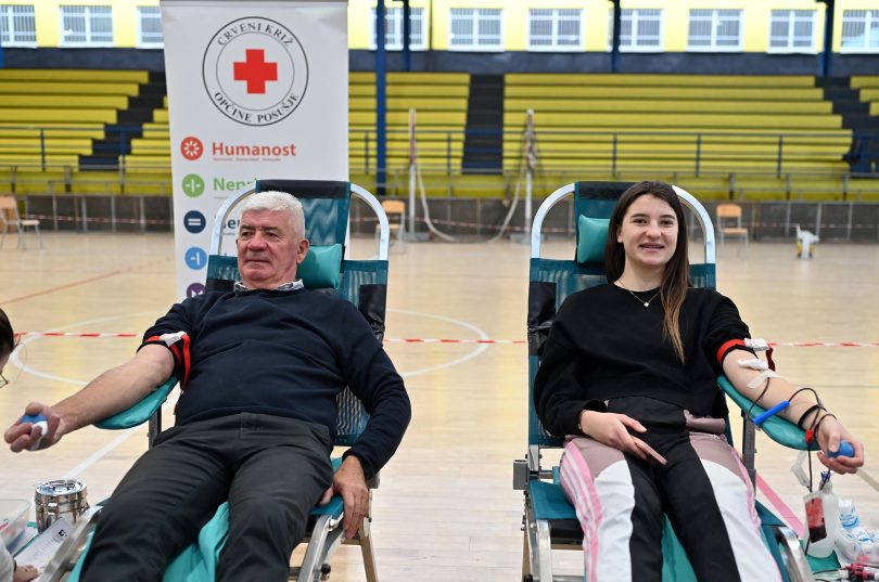 Dva dobrovoljca sjedeće tijekom darivanja krvi u sportskoj dvorani, s logom Crvenog križa u pozadini