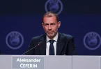 Muškarac u odijelu i kravati sjedu za mikrofonom na konferenciji s UEFA logotipima u pozadini