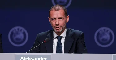 Muškarac u odijelu i kravati sjedu za mikrofonom na konferenciji s UEFA logotipima u pozadini