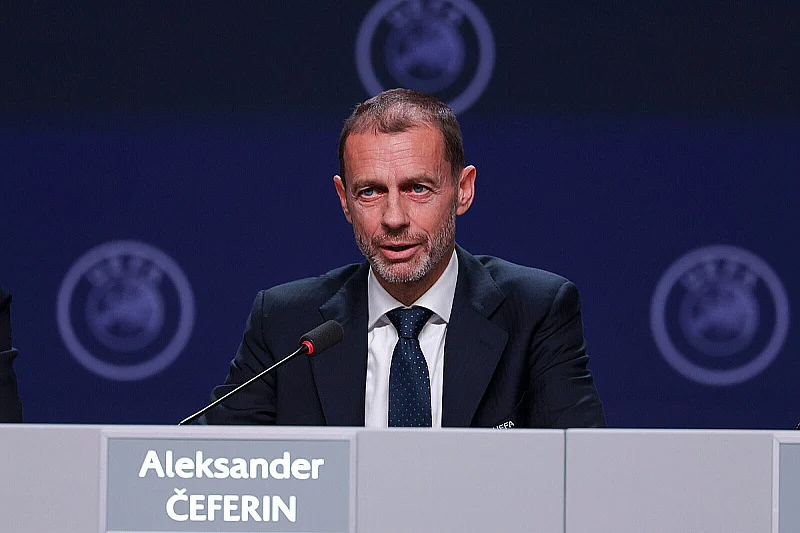 Muškarac u odijelu i kravati sjedu za mikrofonom na konferenciji s UEFA logotipima u pozadini