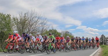 Grupa biciklista u bikelima voze u koloni po asfaltnoj cesti sa zelenilom u pozadini