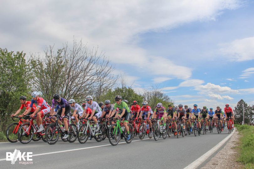 Grupa biciklista u bikelima voze u koloni po asfaltnoj cesti sa zelenilom u pozadini