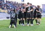 Fudbalski igrači u crnim dresovima u krugu prije utakmice, sa navijačima u pozadini