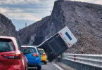 Crveni automobil i prevrnuta bijela prikolica na mostu, stijena u pozadini