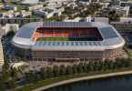 Moderni stadion s crvenim i plavim sjedalima uz obalu rijeke u urbanom okruženju