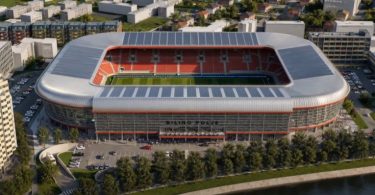 Moderni stadion s crvenim i plavim sjedalima uz obalu rijeke u urbanom okruženju