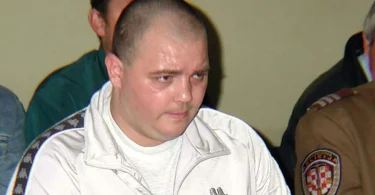 Muško lice tokom sudskog ročišta, u bijeloj majici i tamnoj uniformi u pozadini