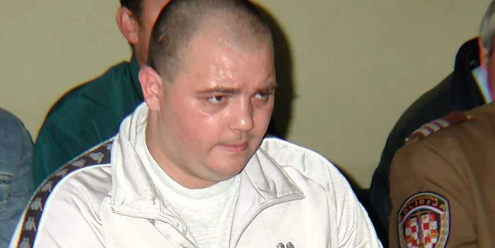 Muško lice tokom sudskog ročišta, u bijeloj majici i tamnoj uniformi u pozadini