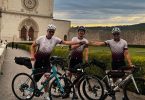 Tri bициклиста u sportskoj opremi ispred crkve, s biciklima spremni za vožnju