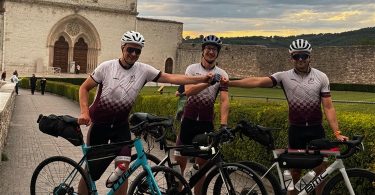 Tri bициклиста u sportskoj opremi ispred crkve, s biciklima spremni za vožnju