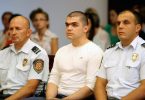 Muž u bijeloj majici između dva pripadnika sigurnosnih snaga tijekom sudskog ročišta