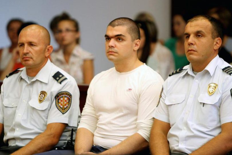 Muž u bijeloj majici između dva pripadnika sigurnosnih snaga tijekom sudskog ročišta