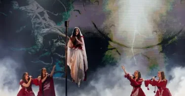 Pjevačica u bijeloj haljini na pozornici okružena pjevačima u crvenim kostimima i dramatičnom scenografijom