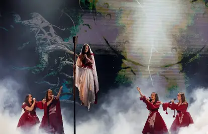 Pjevačica u bijeloj haljini na pozornici okružena pjevačima u crvenim kostimima i dramatičnom scenografijom