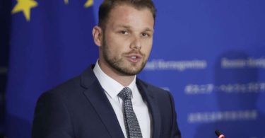 Muškarac u crnom odijelu i kravati govori pred mikrofonom ispred EU zastave