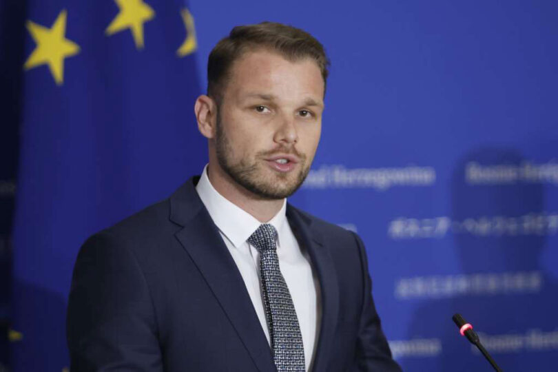 Muškarac u crnom odijelu i kravati govori pred mikrofonom ispred EU zastave